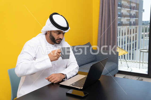استخدام جهاز تقني حديث ومتطور، رجل عربي خليجي اماراتي يرتدي الكندورة والغترة البيضاء يجلس خلف الطاولة يشرب القهوة ويستخدم الحاسوب المحمول، إنجاز مهام العمل عن بعد، إدارة شؤون الأعمال وتنظيمها عبر اللابتوب، الاستمتاع بالمشروبات الساخنة في المنزل