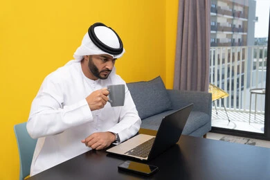 Using a modern and advanced technical device, an Arab Gulf Emirati man wearing a white kandura and ghutra sits behind a table drinking coffee and using a laptop, completing work tasks remotely, managing and organizing business affairs via the laptop, and enjoying hot drinks at home.