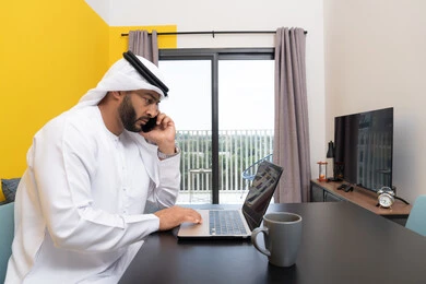 اجراء مكالمة هاتفية عبر الهاتف، العمل عن بعد، انجاز المهام وإدارة شؤون الأعمال وتنظيمها عبر اللابتوب، الاستمتاع بشرب القهوة في المنزل، استخدام جهاز تقني حديث ومتطور، رجل عربي خليجي اماراتي يرتدي الكندورة والغترة يجلس خلف الطاولة ويستخدم الحاسوب المحمول