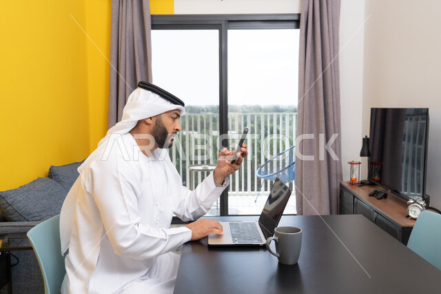 تصفح التطبيقات وبرامج التواصل الاجتماعي عبر الهاتف، إدارة شؤون الأعمال وتنظيمها عبر اللابتوب، الاستمتاع بشرب القهوة في المنزل، استخدام جهاز تقني حديث ومتطور، رجل عربي خليجي اماراتي يرتدي الكندورة والغترة يجلس خلف الطاولة ويستخدم الحاسوب المحمول، العمل عن بعد