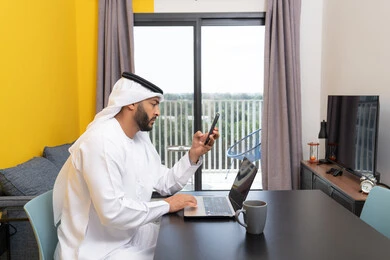 تصفح التطبيقات وبرامج التواصل الاجتماعي عبر الهاتف، إدارة شؤون الأعمال وتنظيمها عبر اللابتوب، الاستمتاع بشرب القهوة في المنزل، استخدام جهاز تقني حديث ومتطور، رجل عربي خليجي اماراتي يرتدي الكندورة والغترة يجلس خلف الطاولة ويستخدم الحاسوب المحمول، العمل عن بعد