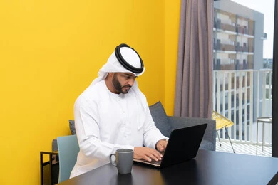 إدارة شؤون الأعمال وتنظيمها عبر اللابتوب، الاستمتاع بالمشروبات الساخنة في المنزل، استخدام جهاز تقني حديث ومتطور، رجل عربي خليجي اماراتي يرتدي الكندورة والغترة البيضاء يجلس خلف الطاولة يشرب القهوة ويستخدم الحاسوب المحمول، إنجاز مهام العمل عن بعد