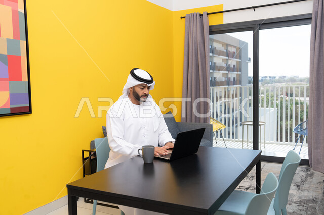 Enjoying hot drinks at home, using a modern and advanced technical device, an Arab Gulf Emirati man wearing a white kandura and ghutra sitting behind a table drinking coffee and using a laptop, completing work tasks remotely, managing and organizing business affairs via the laptop