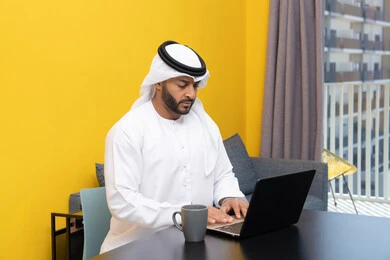استخدام جهاز تقني حديث ومتطور، رجل عربي خليجي اماراتي يرتدي الكندورة والغترة البيضاء يجلس خلف الطاولة يشرب القهوة ويستخدم الحاسوب المحمول، إنجاز مهام العمل عن بعد، إدارة شؤون الأعمال وتنظيمها عبر اللابتوب، الاستمتاع بالمشروبات الساخنة في المنزل