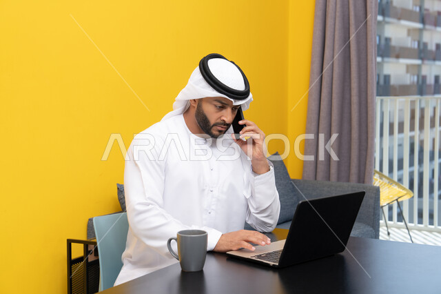 Accomplishing tasks, managing and organizing business affairs via laptop, enjoying drinking coffee at home, using a modern and advanced technical device, an Arab Gulf Emirati man wearing a kandura and ghutra sitting behind a table using a laptop, making a phone call via the phone, working remotely