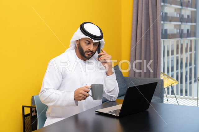 Making a phone call, working remotely, completing tasks, managing and organizing business affairs via laptop, enjoying drinking coffee at home, using a modern and advanced technical device, an Arab Gulf Emirati man wearing a kandura and ghutra sitting behind a table using a laptop