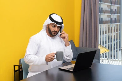 Making a phone call, working remotely, completing tasks, managing and organizing business affairs via laptop, enjoying drinking coffee at home, using a modern and advanced technical device, an Arab Gulf Emirati man wearing a kandura and ghutra sitting behind a table using a laptop