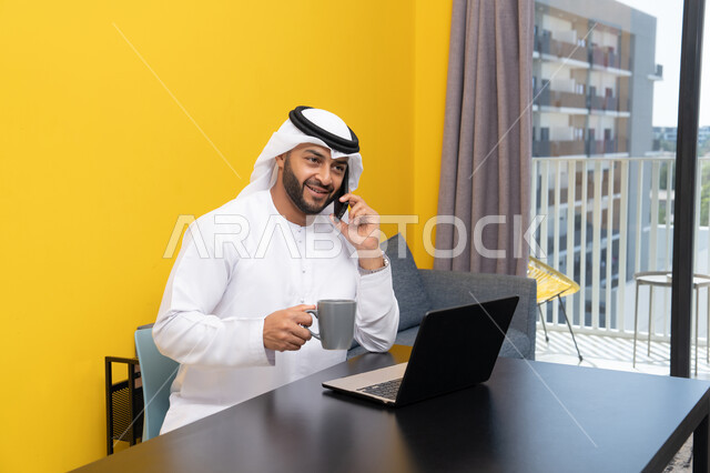 العمل عن بعد، انجاز المهام وإدارة شؤون الأعمال وتنظيمها عبر اللابتوب، الاستمتاع بشرب القهوة في المنزل، استخدام جهاز تقني حديث ومتطور، رجل عربي خليجي اماراتي يرتدي الكندورة والغترة يجلس خلف الطاولة ويستخدم الحاسوب المحمول، اجراء مكالمة هاتفية عبر الهاتف