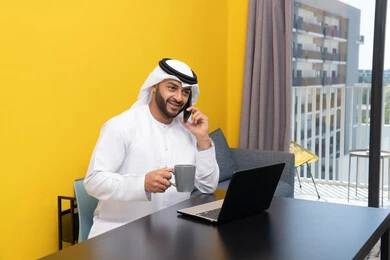 العمل عن بعد، انجاز المهام وإدارة شؤون الأعمال وتنظيمها عبر اللابتوب، الاستمتاع بشرب القهوة في المنزل، استخدام جهاز تقني حديث ومتطور، رجل عربي خليجي اماراتي يرتدي الكندورة والغترة يجلس خلف الطاولة ويستخدم الحاسوب المحمول، اجراء مكالمة هاتفية عبر الهاتف
