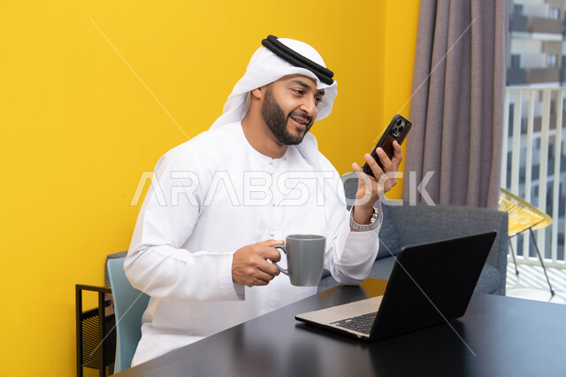 تصفح التطبيقات وبرامج التواصل الاجتماعي عبر الهاتف، إدارة شؤون الأعمال وتنظيمها عبر اللابتوب، الاستمتاع بشرب القهوة في المنزل، استخدام جهاز تقني حديث ومتطور، رجل عربي خليجي اماراتي يرتدي الكندورة والغترة يجلس خلف الطاولة ويستخدم الحاسوب المحمول، العمل عن بعد