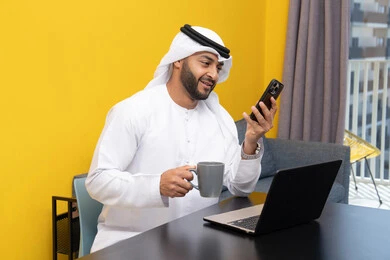 تصفح التطبيقات وبرامج التواصل الاجتماعي عبر الهاتف، إدارة شؤون الأعمال وتنظيمها عبر اللابتوب، الاستمتاع بشرب القهوة في المنزل، استخدام جهاز تقني حديث ومتطور، رجل عربي خليجي اماراتي يرتدي الكندورة والغترة يجلس خلف الطاولة ويستخدم الحاسوب المحمول، العمل عن بعد