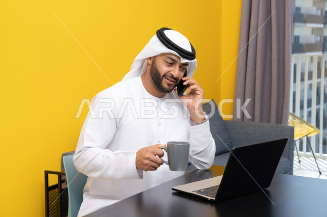 Making a phone call, working remotely, completing tasks, managing and organizing business affairs via laptop, enjoying drinking coffee at home, using a modern and advanced technical device, an Arab Gulf Emirati man wearing a kandura and ghutra sitting behind a table using a laptop