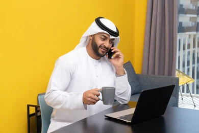 Making a phone call, working remotely, completing tasks, managing and organizing business affairs via laptop, enjoying drinking coffee at home, using a modern and advanced technical device, an Arab Gulf Emirati man wearing a kandura and ghutra sitting behind a table using a laptop