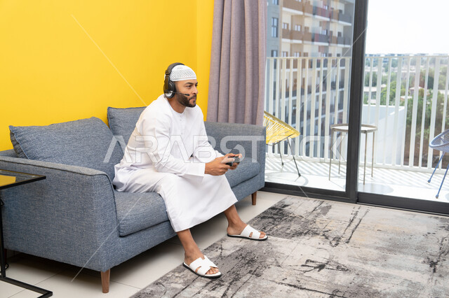 مفهوم الاصغاء والاستمتاع, استخدام الأجهزة التكنولوجية الحديثة، رجل عربي خليجي اماراتي يرتدي الكندورة والقبعة البيضاء يضع سماعات الرأس ويمسك بيده عصا التحكم، قضاء وقت الفراغ باللعب بألعاب الفيديو الترفيهية، الاستماع الى الاغاني عبر الهيدفون