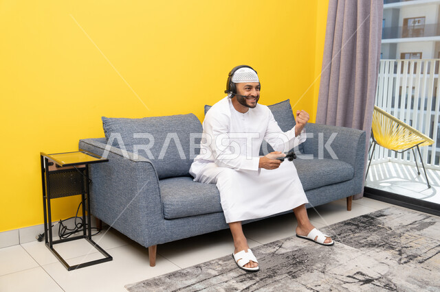 Raising a fist with a victory gesture, using modern technological devices, an Emirati Gulf Arab man wearing a kandura and a white hat, wearing headphones and holding a joystick, spending his free time playing video games, listening to songs on his headphone