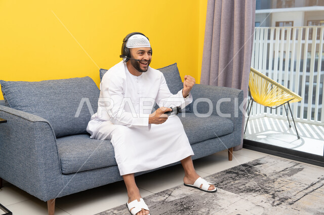الاستماع الى الاغاني عبر الهيدفون، رفع قبضة اليد بايماءات الفوز والنجاح، استخدام الأجهزة التكنولوجية الحديثة، رجل عربي خليجي اماراتي يرتدي الكندورة والقبعة البيضاء يضع سماعات الرأس ويمسك بيده عصا التحكم، قضاء وقت الفراغ باللعب بألعاب الفيديو الترفيهية