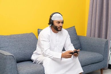The concept of listening and enjoying, using modern technological devices, an Emirati Gulf Arab man wearing a kandura and a white hat, wearing headphones and holding a joystick, spending his free time playing recreational video games, listening to songs through headphones