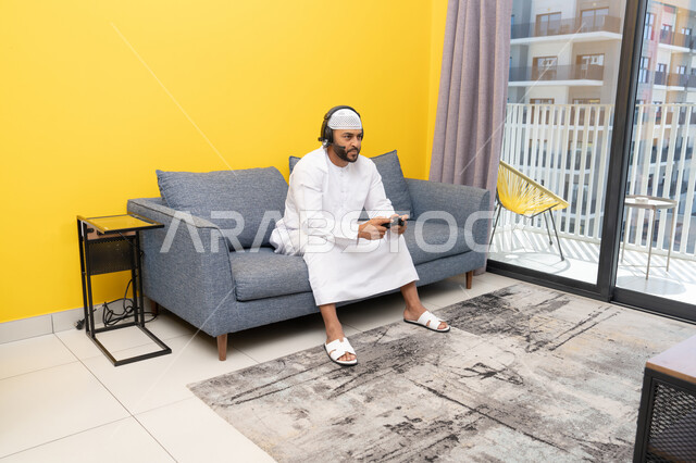 استخدام الأجهزة التكنولوجية الحديثة، رجل عربي خليجي اماراتي يرتدي الكندورة والقبعة البيضاء يضع سماعات الرأس ويمسك بيده عصا التحكم، قضاء وقت الفراغ باللعب بألعاب الفيديو الترفيهية، الاستماع الى الاغاني عبر الهيدفون، مفهوم الاصغاء والاستمتاع