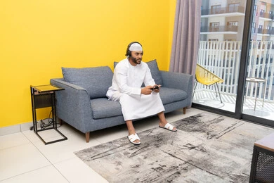 Using modern technological devices, an Emirati Gulf Arab man wearing a kandura and a white hat, wearing headphones and holding a joystick, spending his free time playing recreational video games, listening to songs on headphones, the concept of listening and enjoying