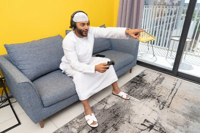 الاشارة باصبع السبابة إلى شيء ما، الاستماع الى الاغاني عبر الهيدفون، استخدام الأجهزة التكنولوجية الحديثة، رجل عربي خليجي اماراتي يرتدي الكندورة والقبعة البيضاء يضع سماعات الرأس ويمسك بيده عصا التحكم، قضاء وقت الفراغ باللعب بألعاب الفيديو الترفيهية