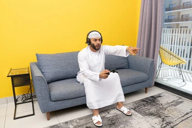 قضاء وقت الفراغ باللعب بألعاب الفيديو الترفيهية، الاشارة باصبع السبابة إلى شيء ما، الاستماع الى الاغاني عبر الهيدفون، استخدام الأجهزة التكنولوجية الحديثة، رجل عربي خليجي اماراتي يرتدي الكندورة والقبعة البيضاء يضع سماعات الرأس ويمسك بيده عصا التحكم