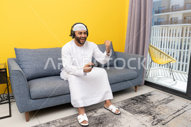 رفع قبضة اليد بايماءات الفوز والنجاح، استخدام الأجهزة التكنولوجية الحديثة، رجل عربي خليجي اماراتي يرتدي الكندورة والقبعة البيضاء يضع سماعات الرأس ويمسك بيده عصا التحكم، قضاء وقت الفراغ باللعب بألعاب الفيديو الترفيهية، الاستماع الى الاغاني عبر الهيدفون