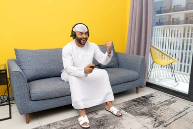 رفع قبضة اليد بايماءات الفوز والنجاح، استخدام الأجهزة التكنولوجية الحديثة، رجل عربي خليجي اماراتي يرتدي الكندورة والقبعة البيضاء يضع سماعات الرأس ويمسك بيده عصا التحكم، قضاء وقت الفراغ باللعب بألعاب الفيديو الترفيهية، الاستماع الى الاغاني عبر الهيدفون