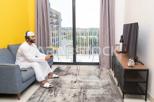 استخدام الأجهزة التكنولوجية الحديثة، رجل عربي خليجي اماراتي يرتدي الكندورة والقبعة البيضاء يضع سماعات الرأس ويمسك بيده عصا التحكم، قضاء وقت الفراغ باللعب بألعاب الفيديو الترفيهية، الاستماع الى الاغاني عبر الهيدفون، مفهوم الاصغاء والاستمتاع