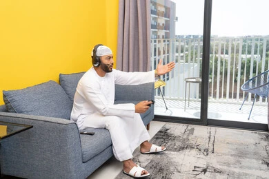 The concept of listening and hearing through headphones, opening the palm of the hand with gestures of sadness and loss, spending free time playing recreational video games, using modern technological devices, an Arab Gulf Emirati man wearing a kandura and a white hat, putting on headphones and holding a joystick in his hand