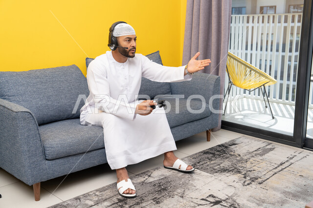 فتح كف اليد بايماءات الحزن والخسارة، قضاء وقت الفراغ باللعب بألعاب الفيديو الترفيهية، الاستماع الى الاغاني عبر الهيدفون، استخدام الأجهزة التكنولوجية الحديثة، رجل عربي خليجي اماراتي يرتدي الكندورة والقبعة البيضاء يضع سماعات الرأس ويمسك بيده عصا التحكم