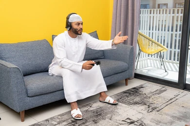 Opening the palm of the hand with gestures of sadness and loss, spending free time playing recreational video games, listening to songs on headphones, using modern technological devices, an Arab Gulf Emirati man wearing a kandura and a white hat, putting on headphones and holding a joystick in his hand