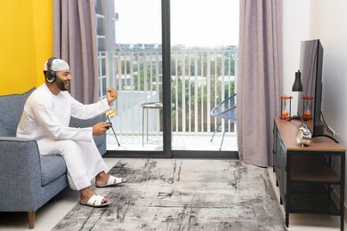 الاستماع الى الاغاني عبر الهيدفون، رفع قبضة اليد بايماءات الفوز والنجاح، استخدام الأجهزة التكنولوجية الحديثة، رجل عربي خليجي اماراتي يرتدي الكندورة والقبعة البيضاء يضع سماعات الرأس ويمسك بيده عصا التحكم، قضاء وقت الفراغ باللعب بألعاب الفيديو الترفيهية