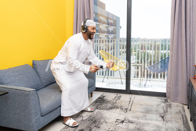 رفع قبضة اليد بايماءات الفوز والنجاح، استخدام الأجهزة التكنولوجية الحديثة، رجل عربي خليجي اماراتي يرتدي الكندورة والقبعة البيضاء يضع سماعات الرأس ويمسك بيده عصا التحكم، قضاء وقت الفراغ باللعب بألعاب الفيديو الترفيهية، الاستماع الى الاغاني عبر الهيدفون