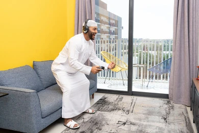 رفع قبضة اليد بايماءات الفوز والنجاح، استخدام الأجهزة التكنولوجية الحديثة، رجل عربي خليجي اماراتي يرتدي الكندورة والقبعة البيضاء يضع سماعات الرأس ويمسك بيده عصا التحكم، قضاء وقت الفراغ باللعب بألعاب الفيديو الترفيهية، الاستماع الى الاغاني عبر الهيدفون