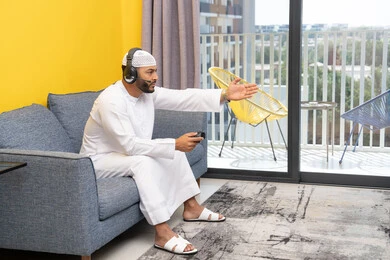 Opening the palm of the hand with gestures of sadness and loss, spending free time playing recreational video games, listening to songs on headphones, using modern technological devices, an Arab Gulf Emirati man wearing a kandura and a white hat, putting on headphones and holding a joystick in his hand