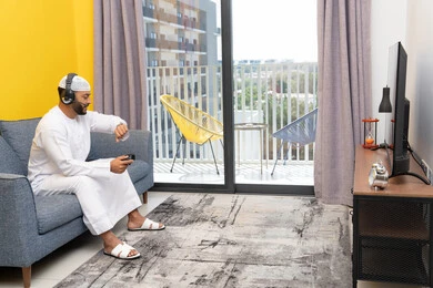 Looking at the time on a wristwatch, the concept of listening and hearing through headphones, spending free time playing recreational video games, using modern technological devices, an Arab Gulf Emirati man wearing a kandura and a white hat, putting on headphones and holding a joystick in his hand