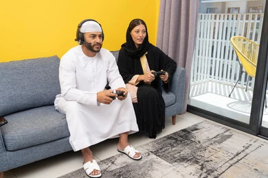 The concept of entertainment, fun and having a good time, listening and hearing through headphones, playing entertainment video games, using modern technological devices, an Emirati Gulf Arab man wearing a kandura and a white hat, putting on headphones and holding a joystick in his hand, sitting with his wife at home