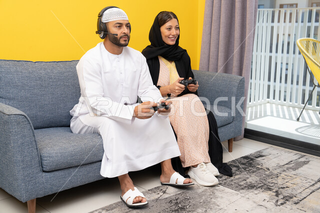 Entertainment, fun, and having a good time, listening to music on headphones, playing video games, using modern technological devices, an Emirati Gulf Arab man wearing a kandura and a white hat, putting on headphones and holding a joystick, sitting with his wife at home