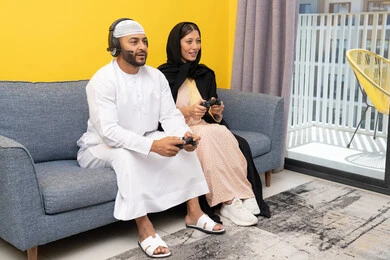 قضاء وقت ممتع، مفهوم التسلية والمتعة، الاصغاء والاستماع عبر الهيدفون، اللعب بألعاب الفيديو الترفيهية، استخدام الأجهزة التكنولوجية الحديثة، رجل عربي خليجي اماراتي يرتدي الكندورة والقبعة البيضاء يضع سماعات الرأس ويمسك بيده عصا التحكم يجلس مع زوجته في المنزل