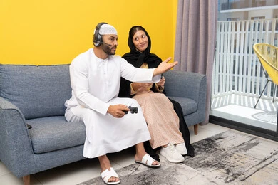 قضاء وقت ممتع، مفهوم التسلية والمتعة، الاصغاء والاستماع عبر الهيدفون، اللعب بألعاب الفيديو الترفيهية، استخدام الأجهزة التكنولوجية الحديثة، رجل عربي خليجي اماراتي يرتدي الكندورة والقبعة البيضاء يضع سماعات الرأس ويمسك بيده عصا التحكم يجلس مع زوجته في المنزل