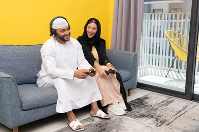 التسلية والمتعة وقضاء وقت ممتع، الاصغاء والاستماع عبر الهيدفون، اللعب بألعاب الفيديو الترفيهية، استخدام الأجهزة التكنولوجية الحديثة، رجل عربي خليجي اماراتي يرتدي الكندورة والقبعة البيضاء يضع سماعات الرأس ويمسك بيده عصا التحكم يجلس مع زوجته في المنزل