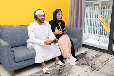 مفهوم التسلية والمتعة وقضاء وقت ممتع، الاصغاء والاستماع عبر الهيدفون، اللعب بألعاب الفيديو الترفيهية، استخدام الأجهزة التكنولوجية الحديثة، رجل عربي خليجي اماراتي يرتدي الكندورة والقبعة البيضاء يضع سماعات الرأس ويمسك بيده عصا التحكم يجلس مع زوجته في المنزل