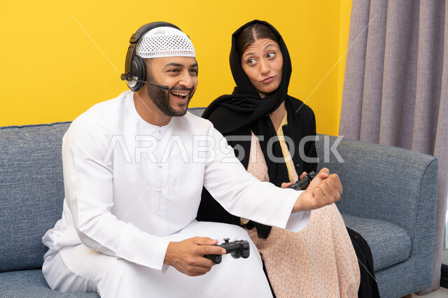 الاستماع والاصغاء عبر الهيدفون، استخدام الأجهزة التكنولوجية الحديثة، رجل عربي خليجي اماراتي يرتدي الكندورة والقبعة البيضاء يضع سماعات الرأس ويمسك بيده عصا التحكم يجلس مع زوجته في المنزل، رفع قبضة اليد بايماءات الحماس والفوز، اللعب بألعاب الفيديو الترفيهية