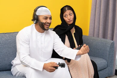 Listening and paying attention through headphones, using modern technological devices, an Arab Gulf Emirati man wearing a kandura and a white hat, wearing headphones and holding a joystick, sitting with his wife at home, raising his fist with gestures of enthusiasm and victory, playing recreational video games
