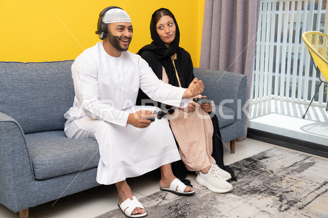 اللعب بألعاب الفيديو الترفيهية، الاصغاء والاستماع عبر الهيدفون، استخدام الأجهزة التكنولوجية الحديثة، رجل عربي خليجي اماراتي يرتدي الكندورة والقبعة البيضاء يضع سماعات الرأس ويمسك بيده عصا التحكم يجلس مع زوجته في المنزل، رفع قبضة اليد بايماءات الحماس والفوز