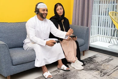 اللعب بألعاب الفيديو الترفيهية، الاصغاء والاستماع عبر الهيدفون، استخدام الأجهزة التكنولوجية الحديثة، رجل عربي خليجي اماراتي يرتدي الكندورة والقبعة البيضاء يضع سماعات الرأس ويمسك بيده عصا التحكم يجلس مع زوجته في المنزل، رفع قبضة اليد بايماءات الحماس والفوز