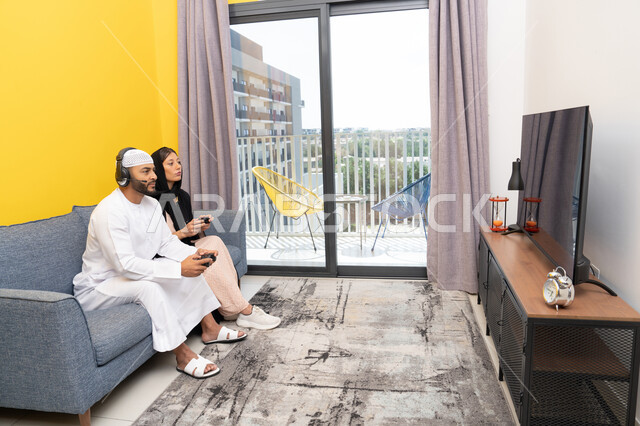 الاصغاء والاستماع عبر الهيدفون، اللعب بألعاب الفيديو الترفيهية، استخدام الأجهزة التكنولوجية الحديثة، رجل عربي خليجي اماراتي يرتدي الكندورة والقبعة البيضاء يضع سماعات الرأس ويمسك بيده عصا التحكم يجلس مع زوجته في المنزل، مفهوم التسلية والمتعة وقضاء وقت ممتع