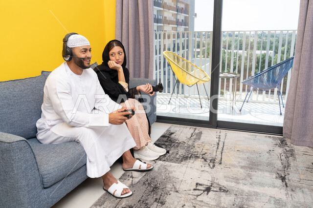 مفهوم المتعة والتسلية، اللعب بألعاب الفيديو الترفيهية، الاصغاء والاستماع عبر الهيدفون، استخدام الأجهزة التكنولوجية الحديثة، رجل عربي خليجي اماراتي يرتدي الكندورة والقبعة يضع سماعات الرأس ويمسك بيده عصا التحكم يجلس مع زوجته في المنزل، ايماءات التركيز والاندماج