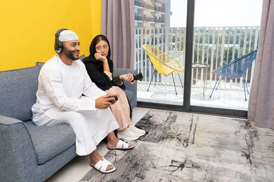 مفهوم المتعة والتسلية، اللعب بألعاب الفيديو الترفيهية، الاصغاء والاستماع عبر الهيدفون، استخدام الأجهزة التكنولوجية الحديثة، رجل عربي خليجي اماراتي يرتدي الكندورة والقبعة يضع سماعات الرأس ويمسك بيده عصا التحكم يجلس مع زوجته في المنزل، ايماءات التركيز والاندماج