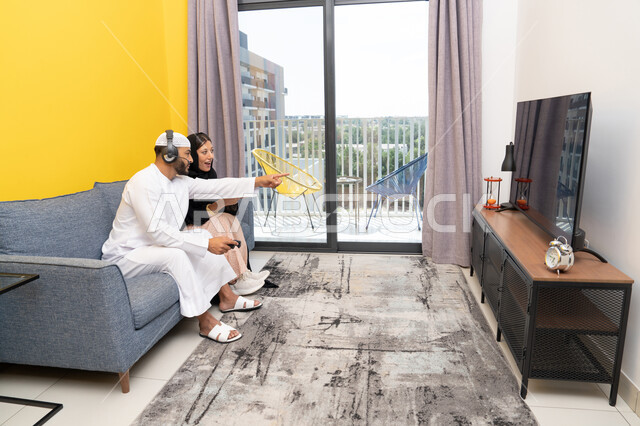 Pointing with the index finger at something, listening and hearing through headphones, playing recreational video games, using modern technological devices, an Arab Gulf Emirati man wearing a kandura and a white hat, putting on headphones and holding a joystick in his hand, sitting with his wife at home